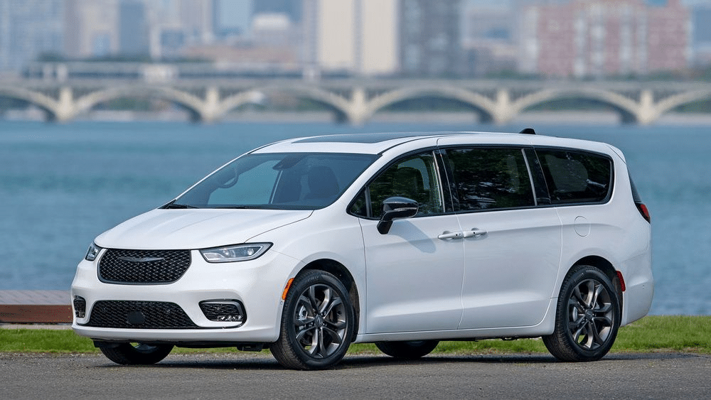 White minivan parked by a waterfront with a cityscape and bridge in the background.