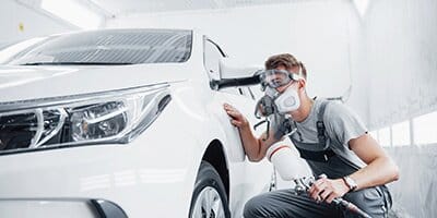 Technician wearing a respirator spray painting a white car in an auto body shop.