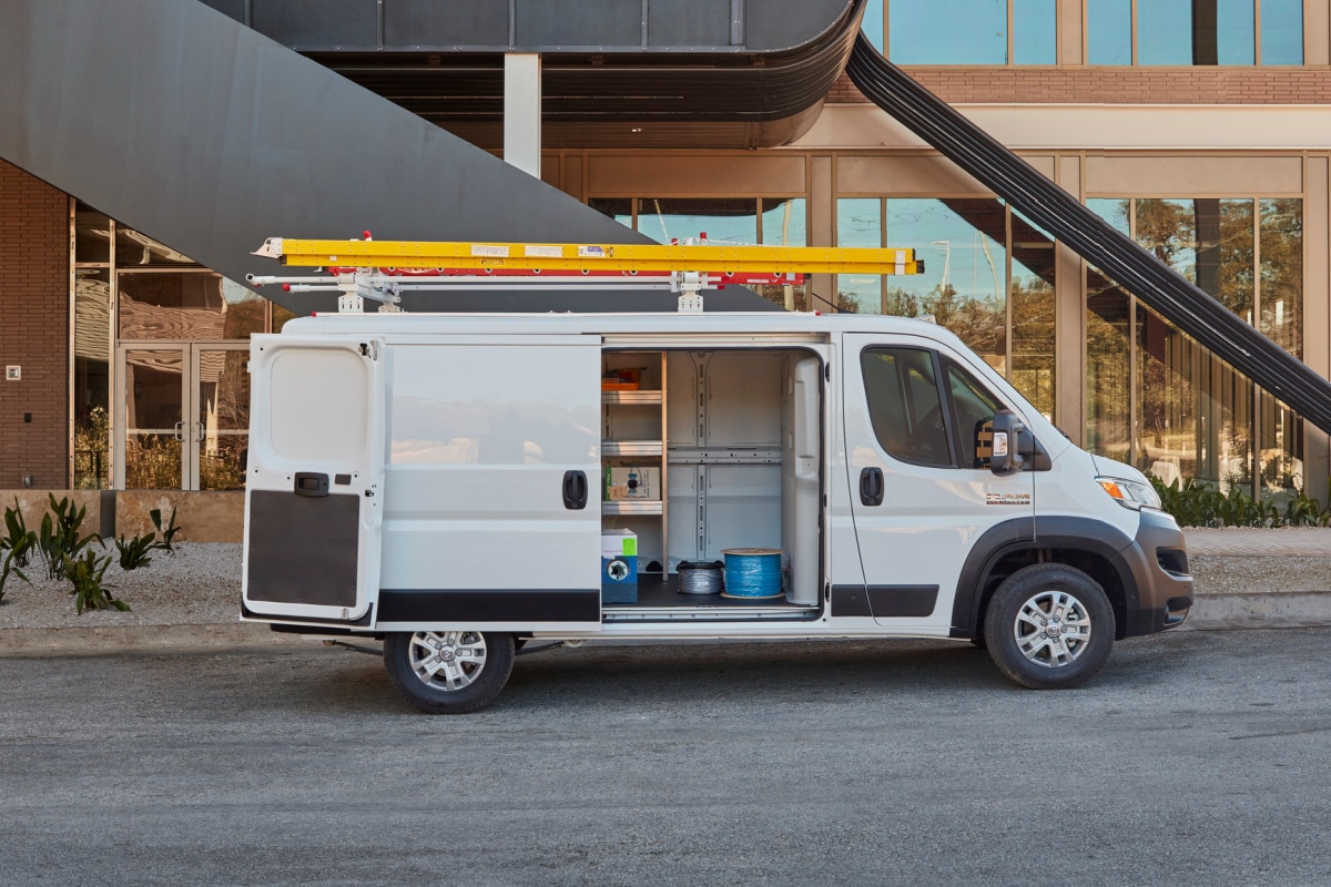 White utility van with open sliding door revealing shelves and equipment, parked outside a modern building.