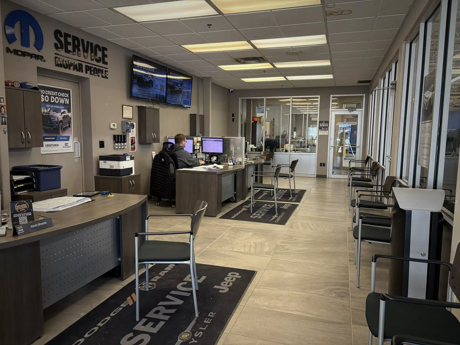 Car dealership service area with desks, chairs, and a staff member at a computer. Mopar signage visible.