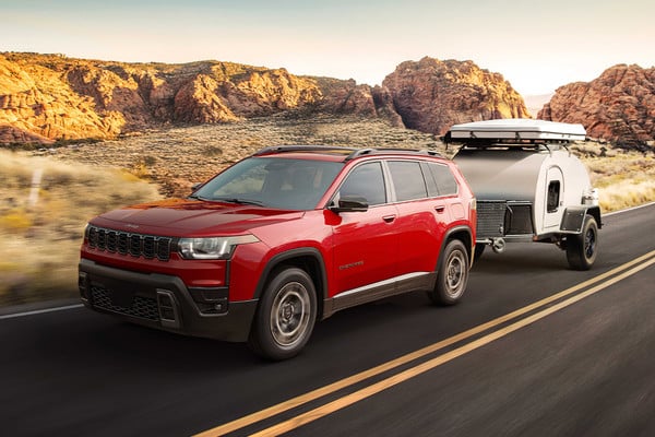 Red SUV towing a camper trailer on a scenic desert road with rocky hills in the background.