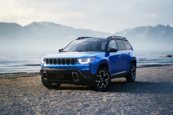 Blue SUV parked on a pebbled beach with mountains and water in the background.